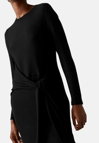 Robe en maille noire à manches longues, présentant un design texturé en côtes et un accent noué à la taille. Silhouette élégante et ajustée.