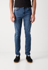 Kék farmernadrág, slim fit szabással, öt zsebes dizájnnal és diszkrét varrással. Fekete tornacipővel párosítva, gumitalppal.