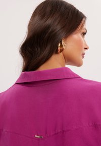 Jasnofuksjowa koszula z kołnierzykiem, charakteryzująca się subtelnym połyskiem oraz złotym akcentem na plecach, prezentująca czysty, strukturalny design.