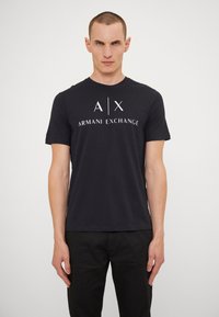 Juodas medvilnės marškinėlis su apykakle, trumpomis rankovėmis ir balta "A|X ARMANI EXCHANGE" logotipu, atspausdintu priekyje.
