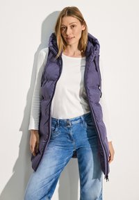 Gilet rembourré violet avec capuche, présentant une fermeture éclair à l'avant, des poches latérales et un design long. En dessous, il y a une chemise blanche à manches longues et un jean bleu.