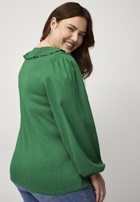 Grøn, struktureret bluse med puffede lange ærmer, har en flæset halsudskæring og en buet kant, båret med blå denimjeans.