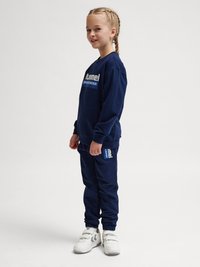 Jong meisje met gevlochten haar, gekleed in marineblauw sportkleding en witte sneakers, staand en glimlachend tegen een effen achtergrond.