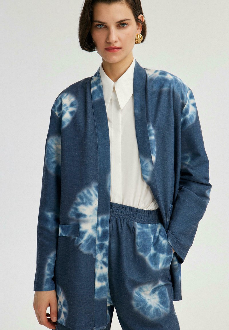Cárdigan azul tie-dye con mangas largas, abertura frontal y dos bolsillos laterales, llevado sobre una camisa blanca con cuello y combinado con pantalones azules.