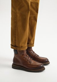 Brązowe skórzane buty za kostkę z lace-up z przodu, teksturowane wykończenie, okrągły czubek i czarny akcent na obcasie. Noszone z podwiniętymi spodniami w kolorze musztardowym.