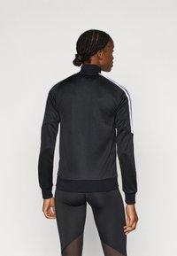 Μαύρο αθλητικό μπουφάν με ψηλό γιακά και λευκές ρίγες στα πλευρά, κατασκευασμένο από λείο, ελαφρύ υλικό. Διαθέτει ριμπ μανσέτες και τελείωμα.