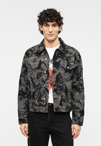 Μαύρο και γκρι τζιν μπουφάν με σχέδιο, τσέπες με καπάκι, κούμπωμα με κουμπιά και γιακάς που ανοίγει, φορεμένο πάνω από λευκό graphic t-shirt.