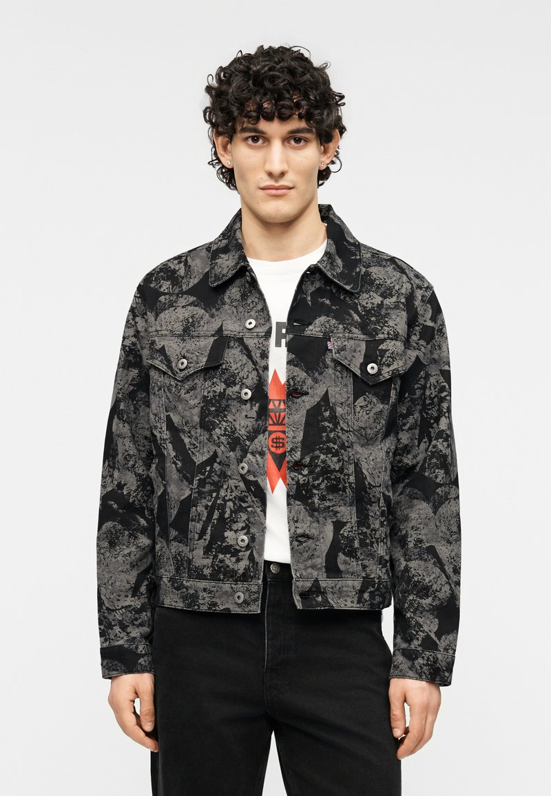 Μαύρο και γκρι τζιν μπουφάν με σχέδιο, τσέπες με καπάκι, κούμπωμα με κουμπιά και γιακάς που ανοίγει, φορεμένο πάνω από λευκό graphic t-shirt.