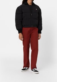 Svart vadderad jacka med dragkedja och ställbar krage, parat med röda raka byxor och svarta sneakers med vita detaljer.