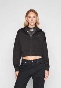 Haut à capuche noir court avec une fermeture éclair à l'avant, doté d'une capuche et d'un logo. Associé à un jean en denim noir taille haute. Texture lisse.