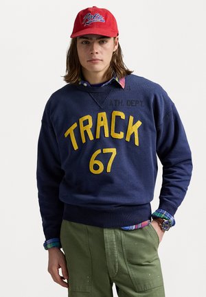 Felpa blu marino con il testo giallo "TRACK 67", abbinata a una camicia a quadri e pantaloni verde oliva. Cappello rosso con logo "Polo" e orologio da polso.