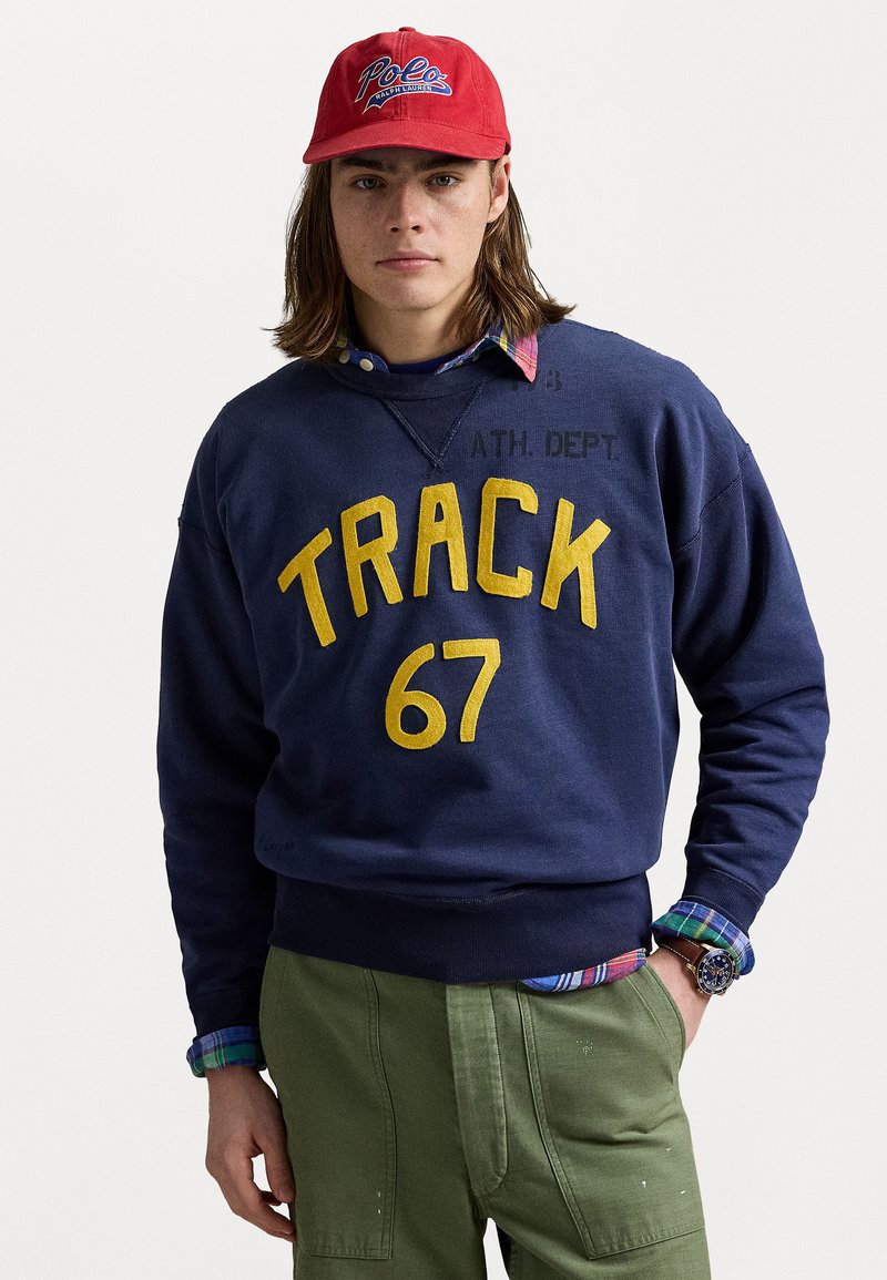Sweatshirt marine avec texte jaune "TRACK 67", associé à une chemise à carreaux et un pantalon olive. Casquette rouge avec logo "Polo" et montre au poignet.