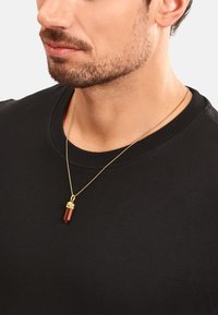 Goldene Halskette mit einem Anhänger aus rotem Kristall, der über einem schwarzen Hemd getragen wird und einen detaillierten Verschluss aufweist. Der Anhänger hat eine geometrische Form und eine glatte Oberfläche.