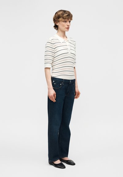 Γυναίκα που φοράει λευκή πλεκτή μπλούζα με κοντά μανίκια και οριζόντια ανοίγματα, σκούρα μπλε τζιν με ίσια μπατζάκια και μαύρα επίπεδα παπούτσια, στέκεται πλαγίως.