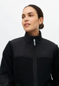 Czarna kurtka polarowa z gładkimi panelami, wysokim kołnierzem, zapięciem na zamek i wytłoczonym logo. Posiada teksturowany materiał na rękawach i klatce piersiowej.