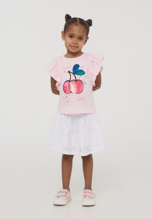 T-shirt rosa con maniche a balze, caratterizzato da un design di ciliegie in paillettes e testo, abbinato a una gonna bianca a balze in pizzo. Sneakers rosa e multicolori.