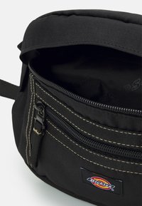 Melnas auduma soma ar rāvējslēdzēja kabatu, ar kontrastējošu šuvumu un logotipa uzšuvumu. Iekšpusē ir plaša nodaļa.