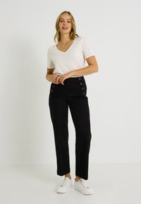 Jeune femme debout portant un haut blanc à manches courtes et col en V, un pantalon noir taille haute avec des boutons, et des baskets compensées blanches.
