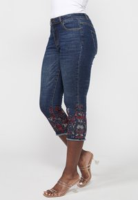 Femme portant un jean court bleu foncé avec une broderie florale rouge et blanche et des sandales à talons transparents, debout de profil.