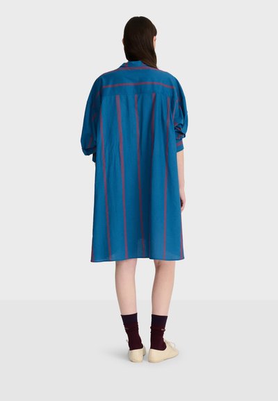 Personne aux longs cheveux foncés portant une robe bleue ample à rayures rouges verticales, des chaussettes foncées et des chaussures beiges, vue de dos sur un fond uni.