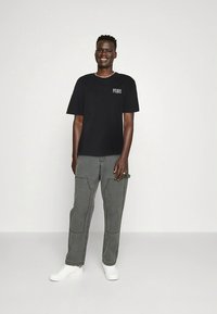 T-shirt en coton noir avec « PEQUS » imprimé sur la poitrine gauche, associé à un pantalon cargo gris et des baskets blanches, sur un fond uni.