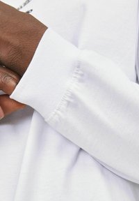 Long-sleeve white cotton shirt with a cuffed sleeve. Fabric has a smooth texture, and the sleeve exhibits slight gathering at the wrist.