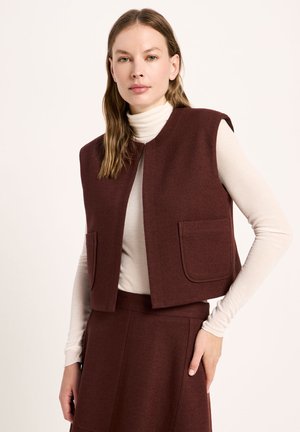 Woman wearing a sleeveless brown vest with front pockets over a white turtleneck and matching brown high-waisted skirt against plain background.