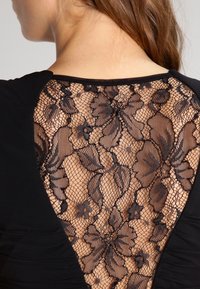 Svart topp med öppen rygg och en blomsterspetsöverdrag. Släta tyg med plisserade detaljer längs sidorna, vilket framhäver texturkontrast.