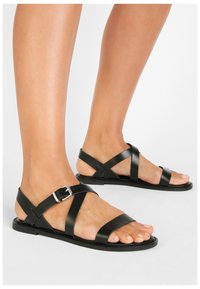 Svarta lädersandaler har flera korsade remmar med spänne. Sålen är platt med minimal textur för komfort.