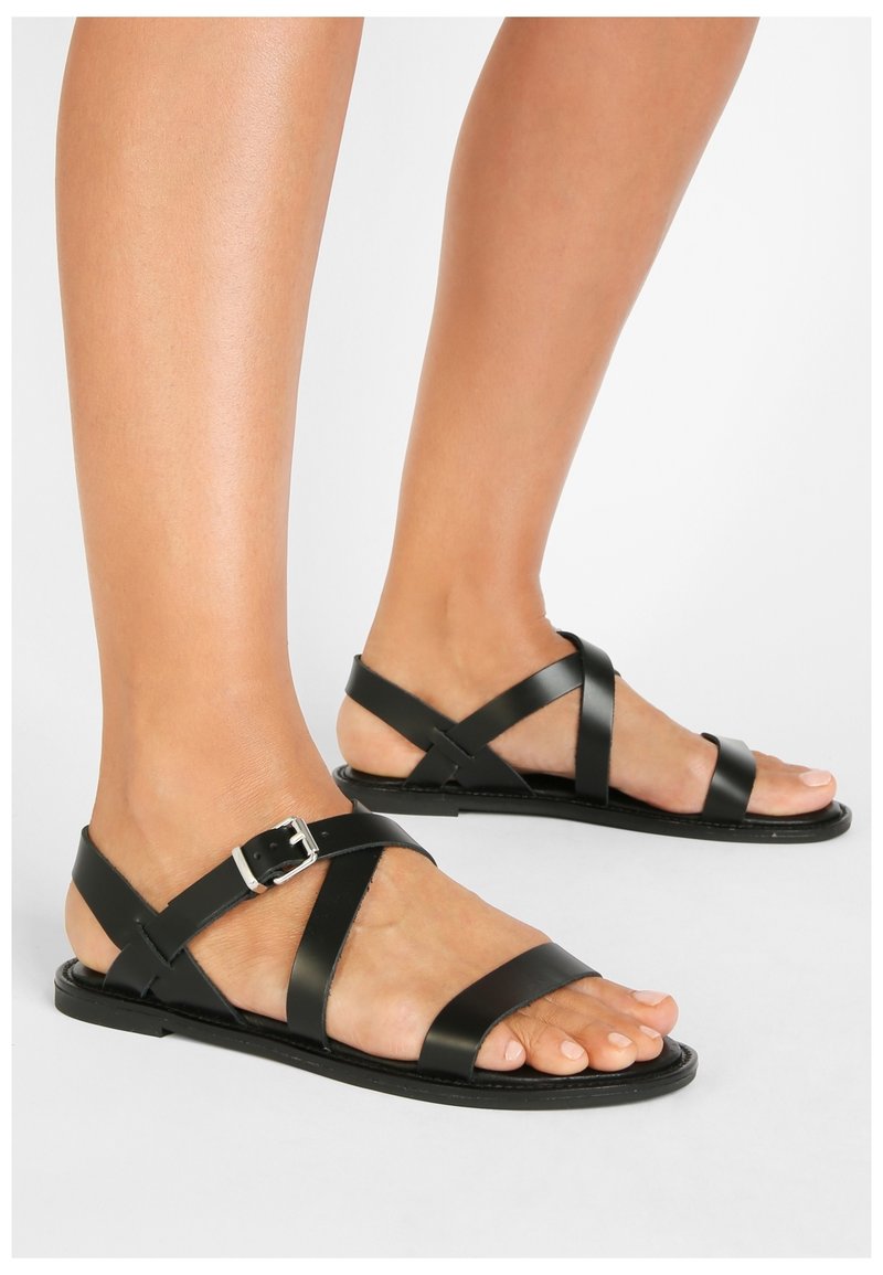 Svarta lädersandaler har flera korsade remmar med spänne. Sålen är platt med minimal textur för komfort.
