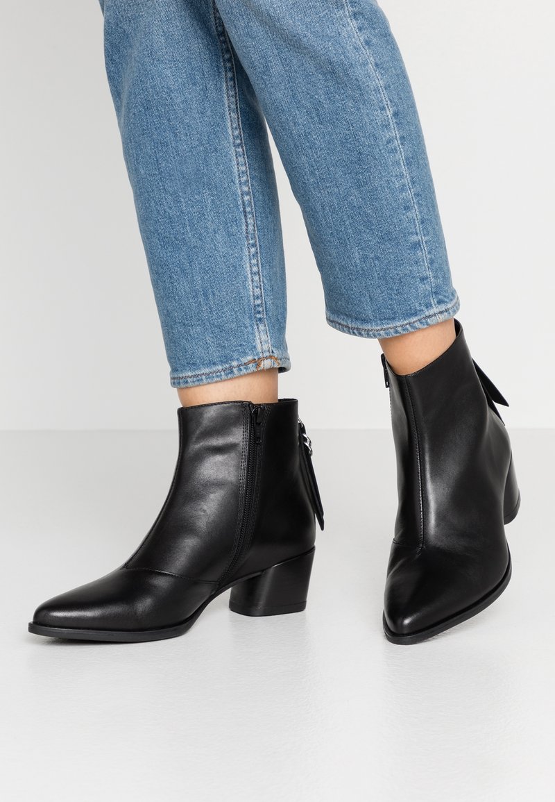 Personne portant un jean bleu coupé et des bottines en cuir noir avec des talons blocs bas et des fermetures éclair latérales.