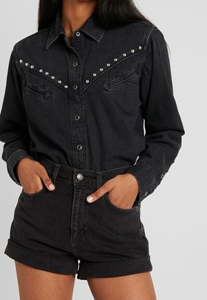 Femme portant une chemise en jean noire à manches longues avec des clous argentés et un short en jean noir assorti aux ourlets retroussés, mains dans les poches.