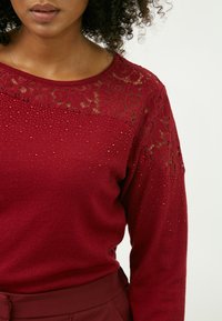 Roter Pullover mit Spitzenakzenten an den Schultern und einer strukturierten Vorderseite mit kleinen dekorativen Perlen, die ein schichtartiges Aussehen erzeugen.