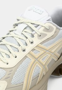 Närbild av en beige och vit Asics löparsko med fokus på meshtyg, snören och logodetaljer på sidan och tungan.