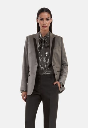 Getextureerde grijze blazer met zwarte revers, gecombineerd met een glanzende sequined zwarte blouse en op maat gemaakte zwarte broeken met knoopdetails.
