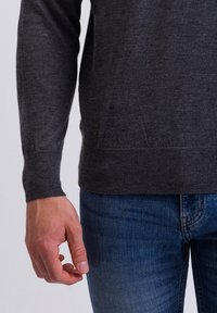 Maglione grigio lavorato a maglia con scollatura rotonda, polsini e orlo a costine, abbinato a jeans in denim blu. Dettaglio di una mano appoggiata di lato.