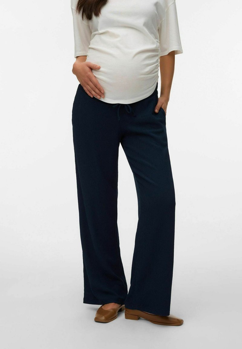 Femme enceinte portant un pantalon ample bleu marine, un haut blanc à manches courtes et des ballerines marron, debout avec une main sur le ventre et l'autre dans la poche.
