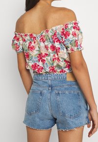 Donna che indossa un top floreale a spalle scoperte con fiori rossi e verdi, abbinato a short di jeans leggeri a vita alta con orli sfilacciati.