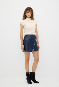 Top aderente beige e una gonna avvolgente in finta pelle blu navy con tasca con zip e dettagli in oro. Stivaletti neri morbidi fino alla caviglia completano il look.
