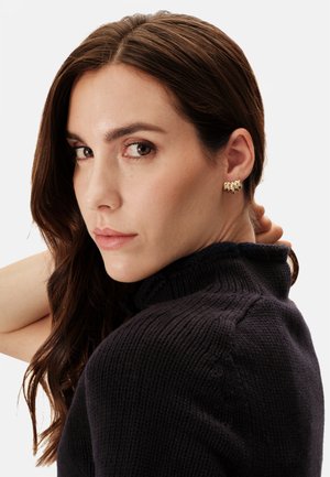 Goldene Creolen mit strukturierter, geschichteter Gestaltung. Das Modell hat lange braune Haare und trägt einen dunklen Strickpullover mit hohem Kragen.