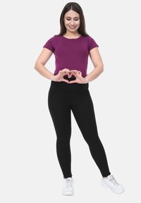Camiseta corta de manga morada combinada con leggings negros, ambos de un diseño ajustado. El modelo forma una figura de corazón con las manos, usando zapatillas blancas.