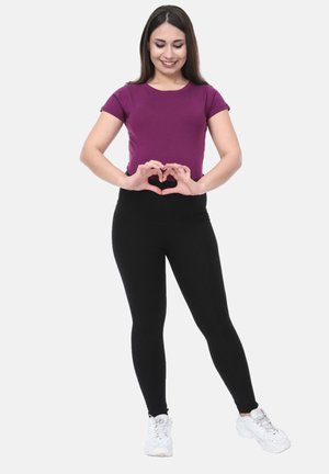 Paarse T-shirt met korte mouwen, gecombineerd met zwarte leggings, beide in een strakke pasvorm. Model vormt een hartvorm met haar handen en draagt witte sneakers.