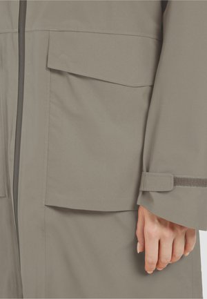 Gros plan sur une poche de veste beige avec rabat, fermeture éclair et sangle réglable au niveau du poignet, portée par une personne dont la main est visible.