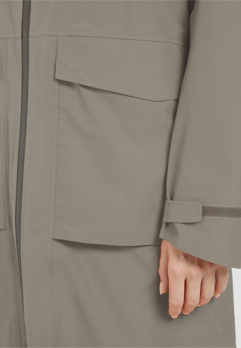 Detailní záběr na kapsu béžové bundy s klopou, zipem a nastavitelným páskem na manžetě, kterou nosí osoba s viditelnou rukou.