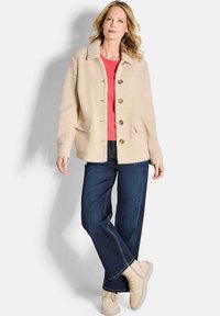 Beige fuzzy jasje met grote knopen, twee voorzakken, gelaagd over een roze shirt, gecombineerd met donkere denim en lichtbeige sneakers.