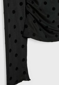 Crni top od tkanine s točkicama koji sadrži reljefne baršunaste crne točkice, glatku teksturu i drapirani dizajn. Rubovi su neuredni.