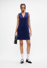 Námornícky kordurovový bezrukávový šaty s V-výstrihom, prednými vreckami a rovnou siluetou. V kombinácii s čiernou kabelkou so cvočkami a bielymi ponožkami.