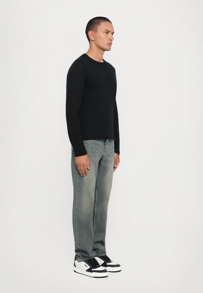 Black long-sleeve shirt paired with faded blue jeans and black-and-white sneakers; model stands sideways against a plain backdrop.
