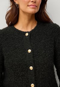 Schwarze, strukturierte Strickjacke mit runder Halslinie, ausgestattet mit vier goldfarbenen Knöpfen und sichtbarem, flauschigem Material. Einfaches Design ohne Muster.