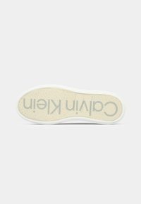 White sneaker sole features a smooth texture with a beige logo print of "Calvin Klein" in a modern font. Subtle pattern along the edges.